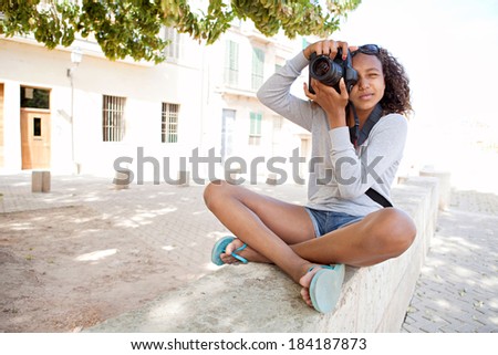 Attractive african american black teenager girl sitting on the bench of a village street, using a slr digital camera taking photographs on a summer holiday. Photography student and tourism lifestyle.