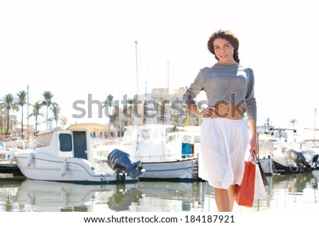 Beautiful young woman walking in a boats and yachts marine pier carrying paper shopping bags and smiling joyfully at the camera during a sunny day on holiday. Travel lifestyle.