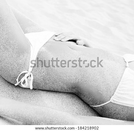 Black and white close up detail of a young attractive woman body laying down relaxing and sunbathing on a white sand beach wearing a white bikini during a summer holiday. Travel and health lifestyle