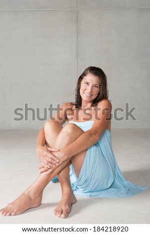 Attractive and elegant mature woman relaxing sitting down on her bathroom floor after a shower, wrapped in a blue cotton towel and smiling at the camera hugging her legs. Beauty lifestyle.