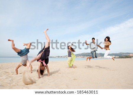 Group of five teenagers friends enjoying a day on the beach together, jumping and doing cartwheels celebrating the summer holidays during a sunny day with a blue sky. Outdoors teenagers lifestyle.