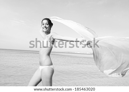 Black and white side view of a young joyful woman on a beach on vacation, holding up a floating silk fabric sarong flying in the breeze against a sunny sky and the sea. Beauty and lifestyle.