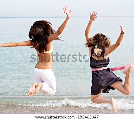 Rear view of two joyful teenager girls friends jumping up in the air together on the shore of a summer beach on holiday during a sunny day. Tourism and travel youth lifestyle.