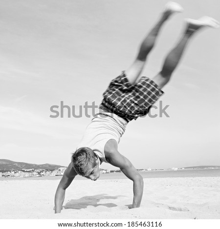 Black and white rear view of a joyful and energetic teenager boy doing cartwheels with his legs up in the air while visiting a summer beach on holiday during a sunny day. Travel youth lifestyle.