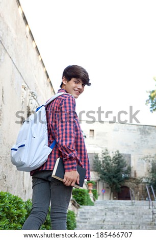Rear portrait of a teenager student boy walking up the stairs in a college campus stone building carrying a text book and a backpack, turning to look at the camera. Student outdoors lifestyle.