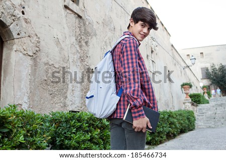 Rear portrait of a teenager student boy walking up the stairs in a college campus stone building carrying a text book and a backpack, turning to look at the camera. Student outdoors lifestyle.