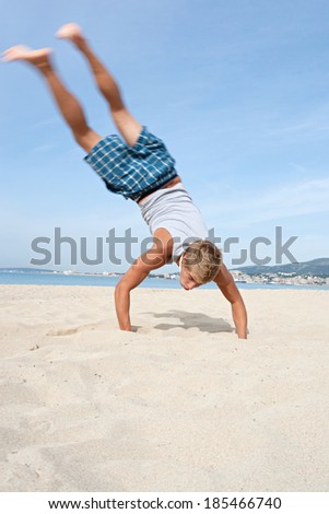 Rear view of a joyful and energetic teenager boy doing cartwheels with his legs up in the air while visiting a summer beach on holiday during a sunny day. Tourism and travel youth lifestyle.