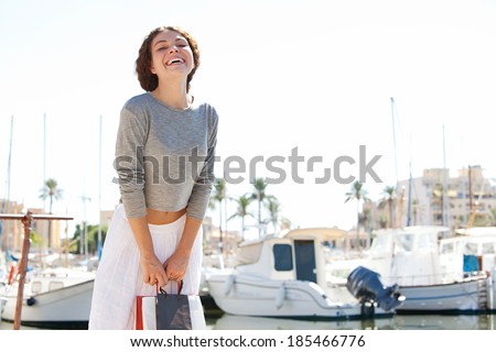 Joyful and attractive young woman standing on a boats marine holding shopping carrier bags while laughing looking at the camera against a sunny blue sky. Travel and healthy lifestyle.