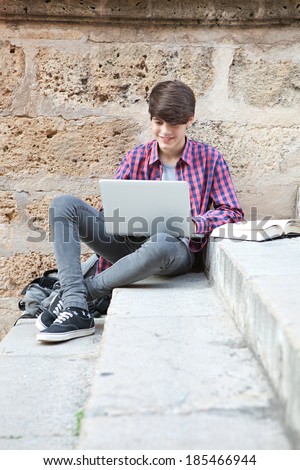 Attractive teenager boy sitting on a college campus with school books and a laptop computer against an old stone building wall doing his homework. Education technology lifestyle.