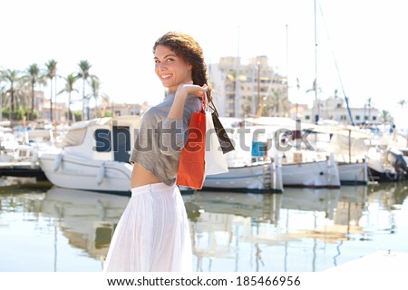 Side view of a beautiful young woman walking in a boats marine pier carrying paper shopping bags and smiling joyfully at the camera during a sunny day on holiday. Travel lifestyle.