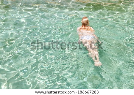 Rear view of a young woman swimming in the clear and transparent blue waters of a health spa swimming pool, relaxing on a summer holiday trip and enjoying a swim during a sunny day, outdoors.