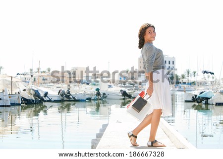 Beautiful young woman joyfully walking with shopping bags in a yachts port while on a holiday trip in a destination city, smiling during a sunny summer day. Consumerism and luxurious travel lifestyle.