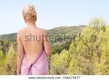 Rear beauty view of an attractive young blond woman back relaxing breathing fresh air in the mountains on vacation, covering her body with a pink silk fabric. Beauty and lifestyle in nature.