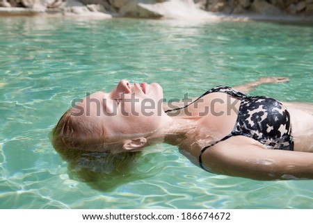 Profile beauty portrait of an attractive scandinavian blond woman floating in a swimming pool of blue transparent water on holiday during a sunny summer day. Travel and vacation lifestyle outdoors.