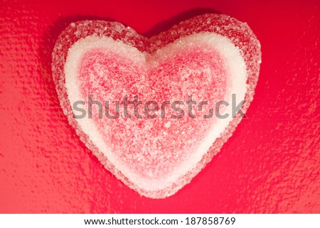 Over head still life close up detail view of a heart shape sugar candy sweet covered in grains of sugar against a bright red background. Interior unhealthy foods and indulgence.