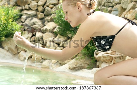 Close up side view of an attractive young blond woman kneeling by a natural swimming pool in a health spa, with water in her hands filtering through her fingers on a summer holiday. Healthy lifestyle.