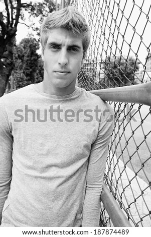Black and white portrait of an attractive teenager young man standing by a college campus building and sports ground with wire fencing, thoughtful during a break. Student outdoors lifestyle.