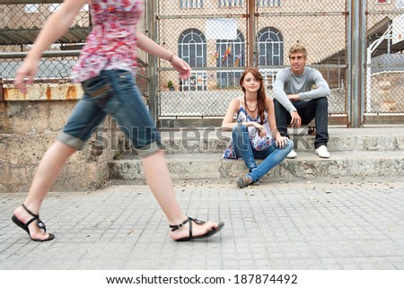 Two teenagers relaxing in a college campus with another young woman body figure walking passed the building. Students outdoors lifestyle.