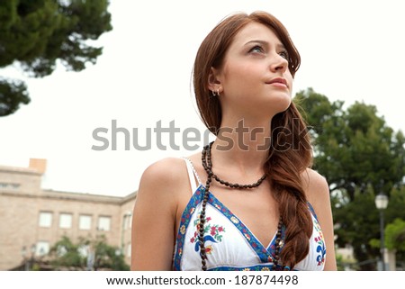 Close up portrait of an attractive student teenager girl standing in a park during a summer day, thoughtfully looking up to the sky. Student outdoors lifestyle.