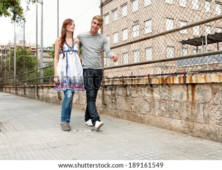 Attractive teenagers boyfriend and girlfriend walking together passed a college university building and sports ground holding each other during a weekend break in an urban city. Youth lifestyle.