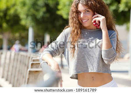 Portrait of a young professional woman using a smartphone to make a call, smiling in the city and leaning on a banister with healthy green trees in the background. Technology lifestyle outdoors.