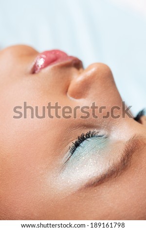 Close up portrait view of an attractive african american black woman laying down against a blue background with her eyes closed wearing fantasy glitter make up cosmetics. Beauty and fashion lifestyle.