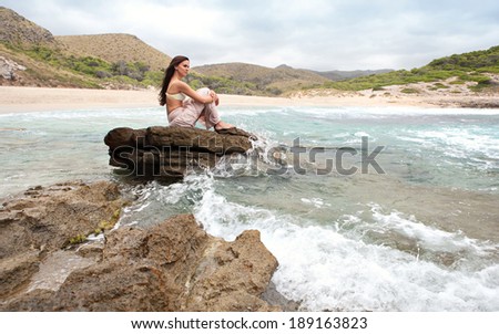 Side view of an attractive young woman sitting and relaxing standing on a rock by the sea, breathing fresh air on a beach and enjoying nature. Health and beauty outdoors lifestyle.