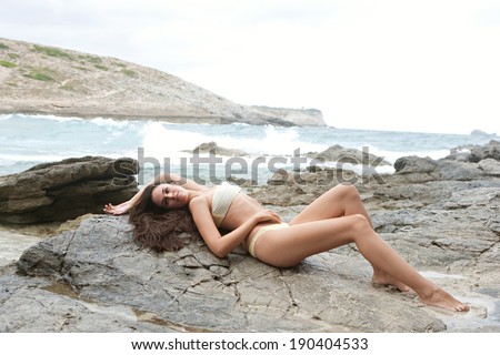 Side body view of a beautiful young woman relaxing laying down on the dark textured rocks of a beach with mountains, lounging and enjoying sunbathing on a vacation by the sea. Beauty and lifestyle.