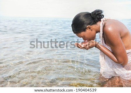Side view of an attractive african american black woman kneeling on the shore of a beach splashing sea water on her face wearing a wet sarong during a summer holiday. Beauty and lifestyle outdoors.