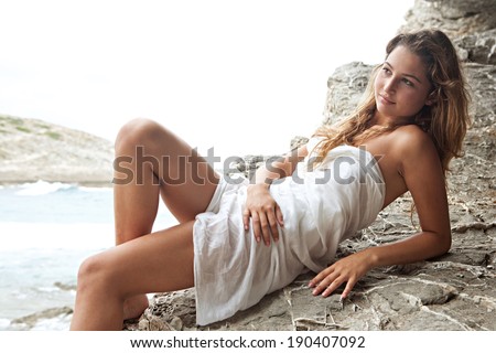 Attractive thoughtful woman relaxing on a coastal dark and textured rock mountain wrapped in a white sarong, sunbathing and relaxing during a summer holiday by the sea. Beauty and health lifestyle.
