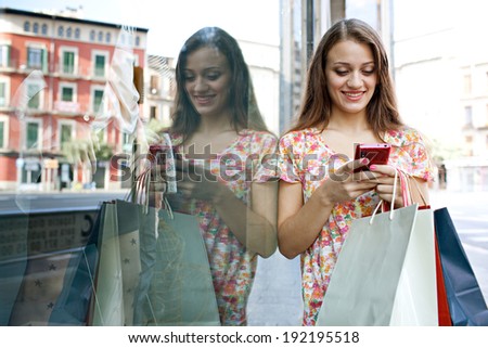 Portrait of an attractive joyful young woman shopping in a city by a fashion store window with reflections, using a smartphone device to network outdoors. Consumerism and technology lifestyle.