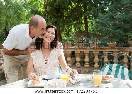 Senior couple sitting together having breakfast in a luxury hotel garden during a sunny day on holiday. Mature people eating and drinking healthy food and kissing, smiling. Outdoors lifestyle.