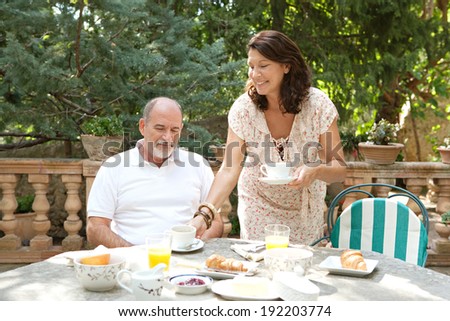 Attractive mature tourists couple having a luxurious continental breakfast in a villa garden table on a sunny day on holiday. Senior people eating healthy food and enjoying life, outdoors lifestyle.