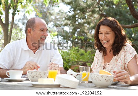 Senior couple having breakfast together at a table in a luxury hotel garden on a sunny day. Mature people eating healthy food and laughing having fun, drinking coffee and juice. Outdoors lifestyle.
