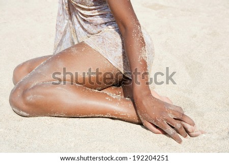 Close up detail view of a beautiful young black woman lower legs section relaxing sunbathing on a beach with white sand grains on her dark tanned skin on vacation. Travel, healthy holiday lifestyle.