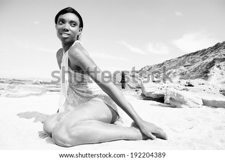 Side view of an attractive young black woman with a perfect body and skin, sitting on a white sand beach during a summer holiday, relaxing sunbathing on a sunny day outdoors. Lifestyle and travel.