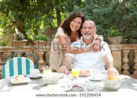 Senior couple sitting together having breakfast in a luxury hotel garden during a sunny day on holiday. Mature people eating and drinking healthy food hugging and smiling. Outdoors lifestyle.
