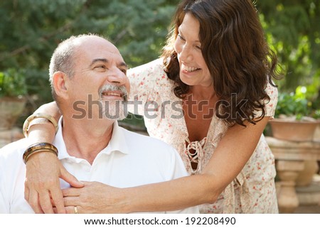 Attractive mature tourists couple sitting in a green garden during a sunny day on a summer holiday enjoying each others company, hugging and loving outdoors. Joyful senior people travel and lifestyle.