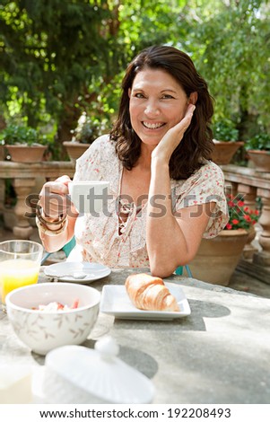 Attractive senior woman at a table having breakfast in a luxury hotel garden during a sunny day on holiday. Mature people eating and drinking healthy food and relaxing, smiling. Outdoors lifestyle.