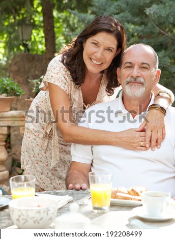 Mature tourists couple having a luxurious continental breakfast in a hotel garden on a sunny day on holiday. Senior people eating healthy food and enjoying each others company, hugging. Lifestyle.