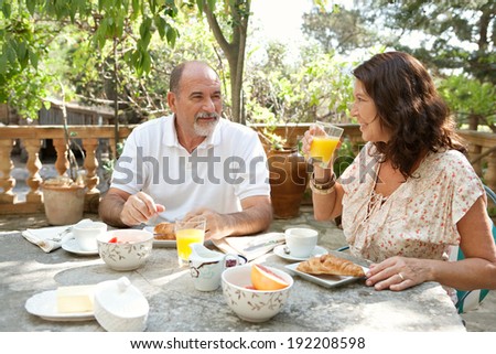 Senior couple having breakfast together at a table in a luxury hotel garden during a sunny day. Mature people eating healthy food and having a conversation. Outdoors lifestyle.