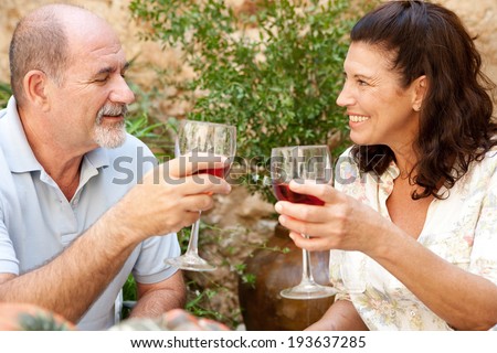 Side portrait of a senior couple relaxing in the garden of a luxury home on holiday, enjoying a glass of wine and toasting. Mature people drinking alcohol sitting at a table. Outdoors lifestyle.