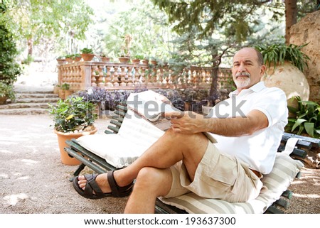 Senior man relaxing in a luxury holiday villa green garden, sitting on a bench and reading a newspaper while on vacation in a Mediterranean countryside hotel. Mature travel and outdoors lifestyle.