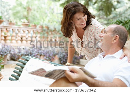 Senior couple husband and wife having a conversation and enjoying a holiday in a luxury hotel garden during a sunny day with the man reading a newspaper. Mature people lifestyle and retirement.
