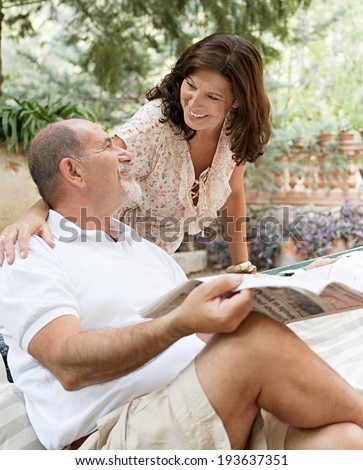 Senior couple husband and wife having a conversation and enjoying a holiday in a luxury hotel garden during a sunny day with the man reading a newspaper. Mature people lifestyle and retirement.