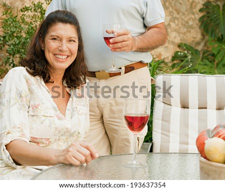Senior couple husband and wife smiling joyfully and enjoying a holiday in a luxury home garden during a sunny day drinking a glass of rose wine. Mature people outdoors lifestyle and retirement.