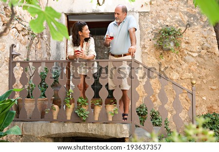 Attractive mature couple on a wooden staircase in a luxury hotel holiday villa green garden on vacation, drinking a glass of wine together and smiling having a conversation. Outdoors senior lifestyle.