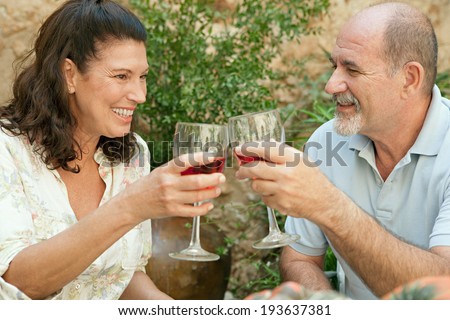 Side portrait of a senior couple relaxing in the garden of a luxury home on holiday, enjoying a glass of wine and toasting. Mature people drinking alcohol sitting at a table. Outdoors lifestyle.