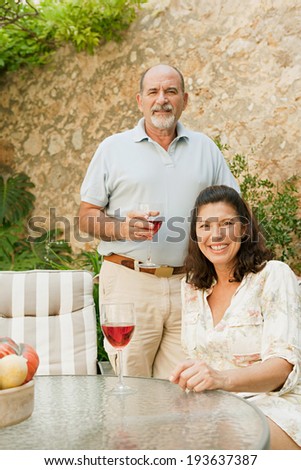 Senior couple husband and wife smiling joyfully and enjoying a holiday in a luxury home garden during a sunny day drinking a glass of rose wine. Mature people outdoors lifestyle and retirement.
