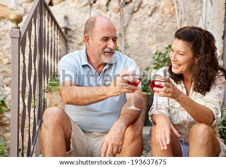 Portrait of a senior man and woman sitting on the stone steps of luxury hotel garden on holiday drinking wine and toasting in celebration, relaxing on vacation. Mature people, outdoors lifestyle.